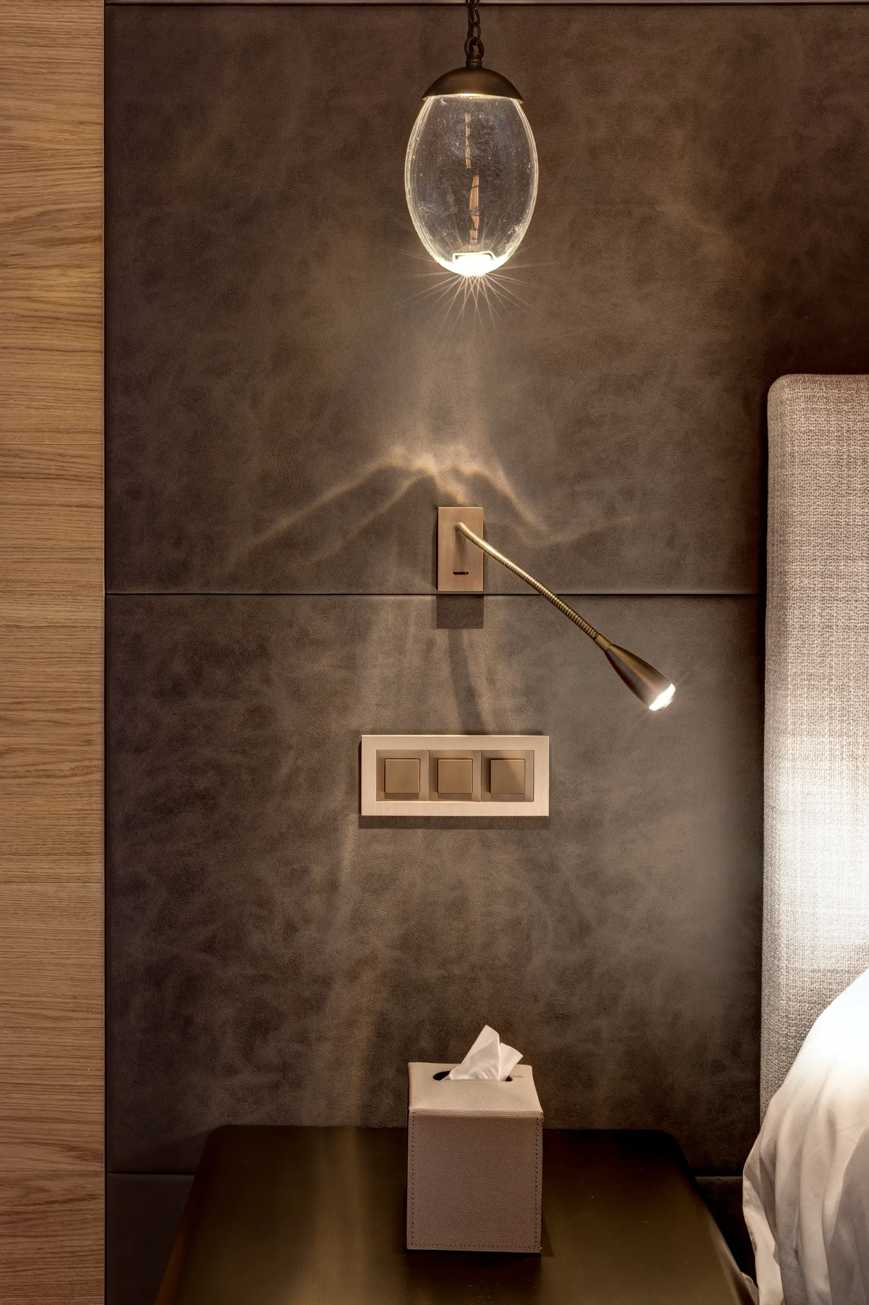 Bedside detail with a glass pendant light and brass reading lamp casting soft pools of light across a leather-clad headboard wall