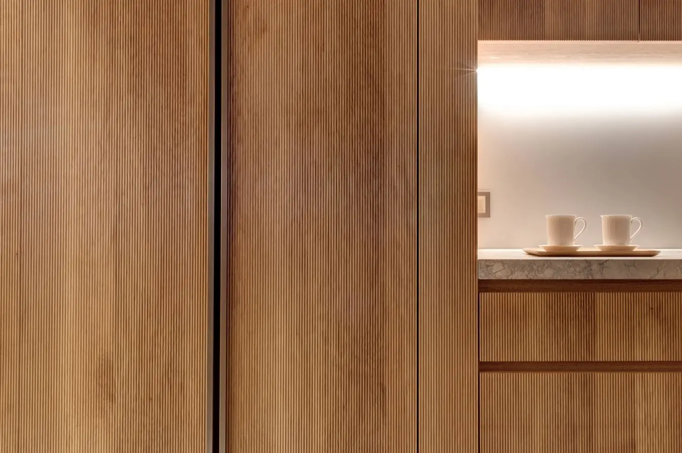 Close-up of fluted oak joinery beside a backlit niche with a marble shelf holding two ceramic cups on a tray