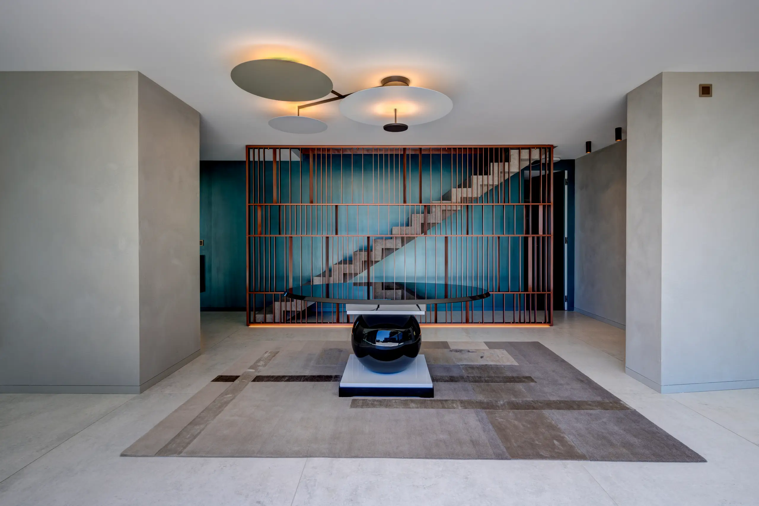 Entrance hall with a concrete staircase rising behind a copper-toned timber screen, three disc pendant lights overhead and a glossy black spherical console on a geometric rug