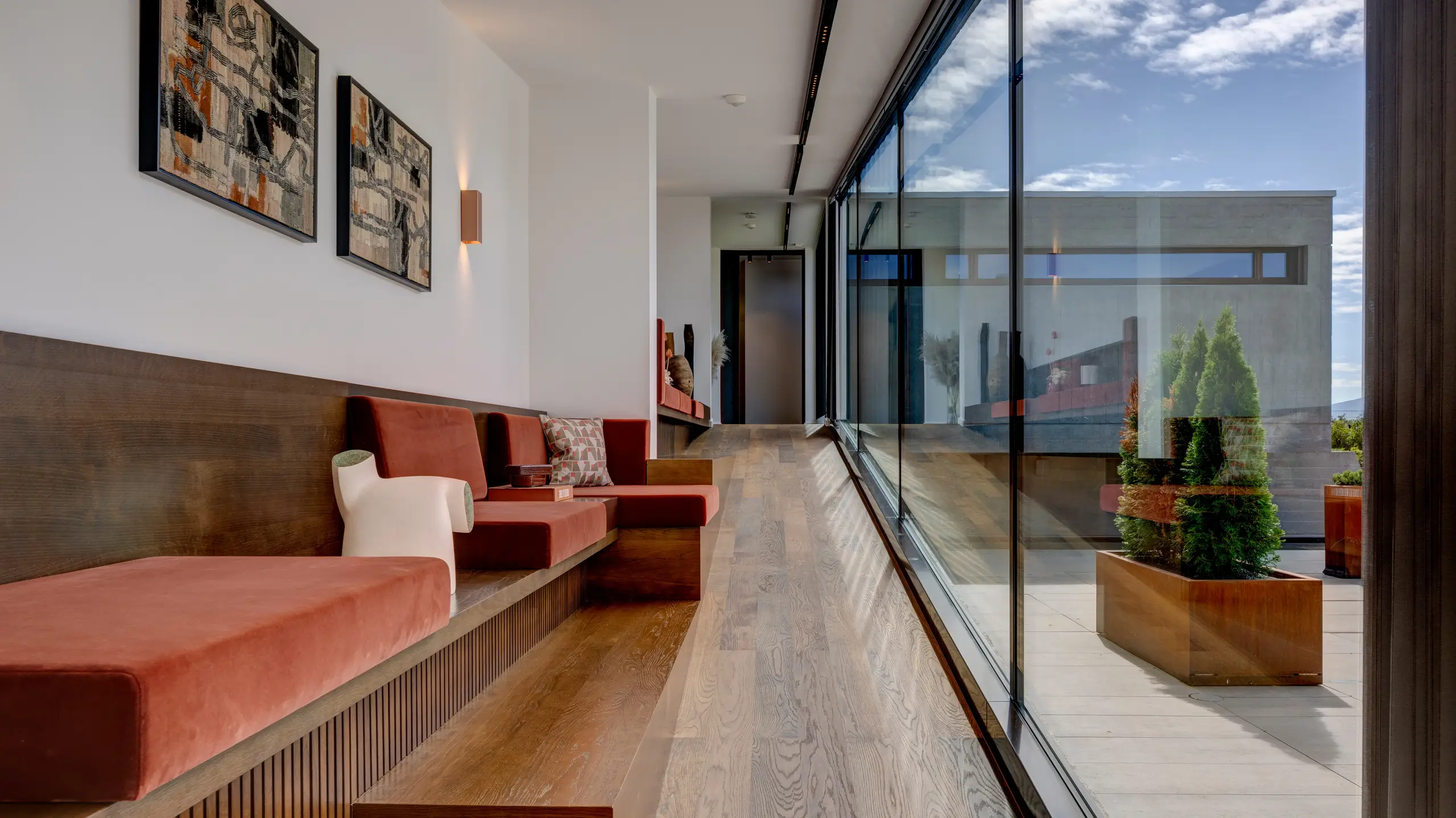 Glazed corridor lined with a built-in banquette in rust-red cushions, opening through a full-height glass wall to the garden