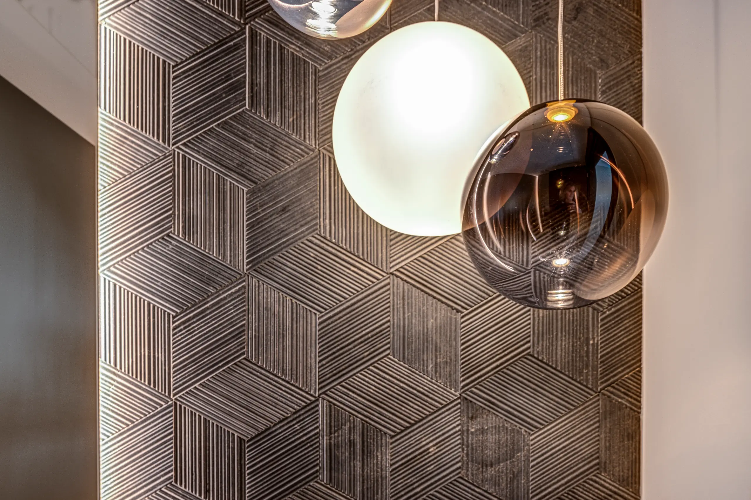 Detail of three spherical pendant lights — frosted, smoked and mirror-finished — against a patterned tiled wall