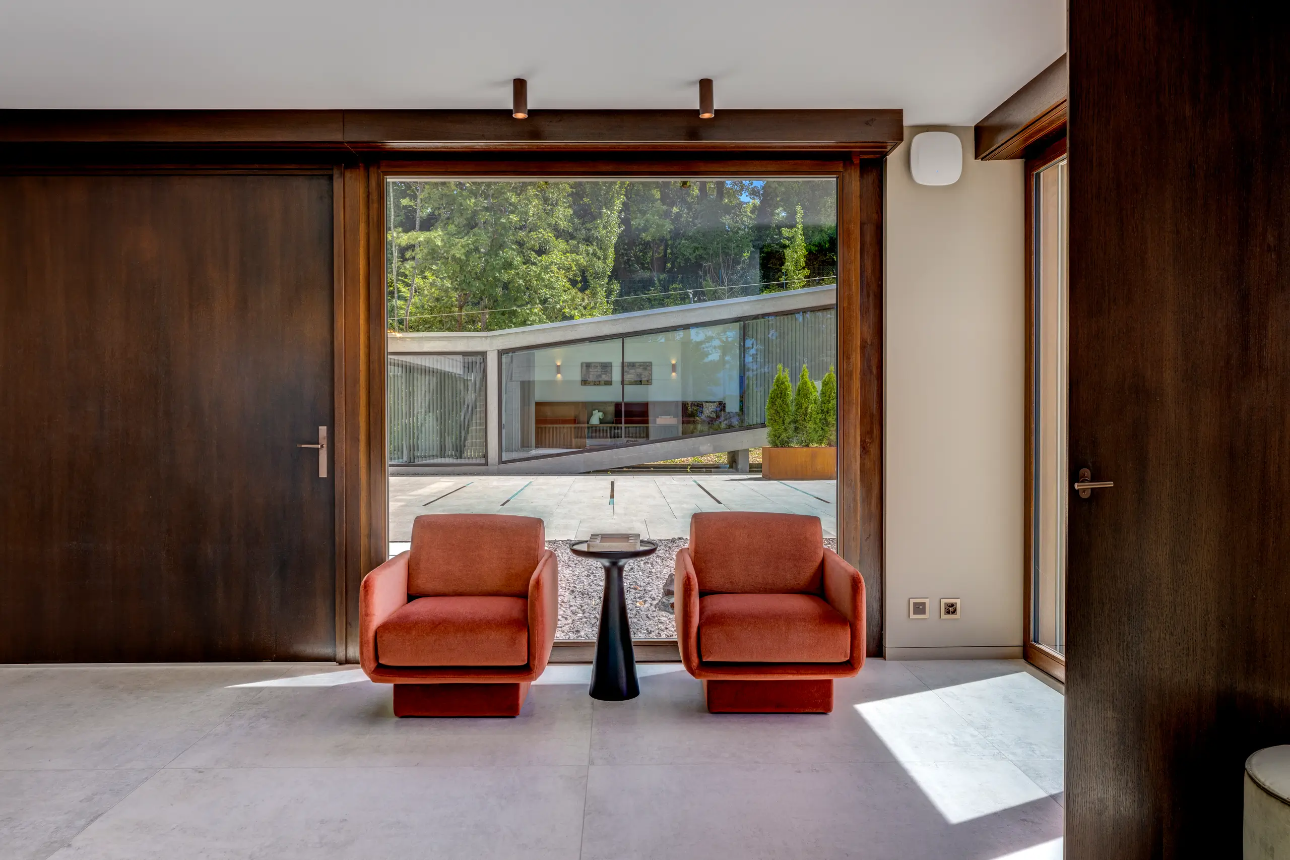 Timber-framed room with two rust-orange armchairs and a black pedestal table beneath ribbon windows