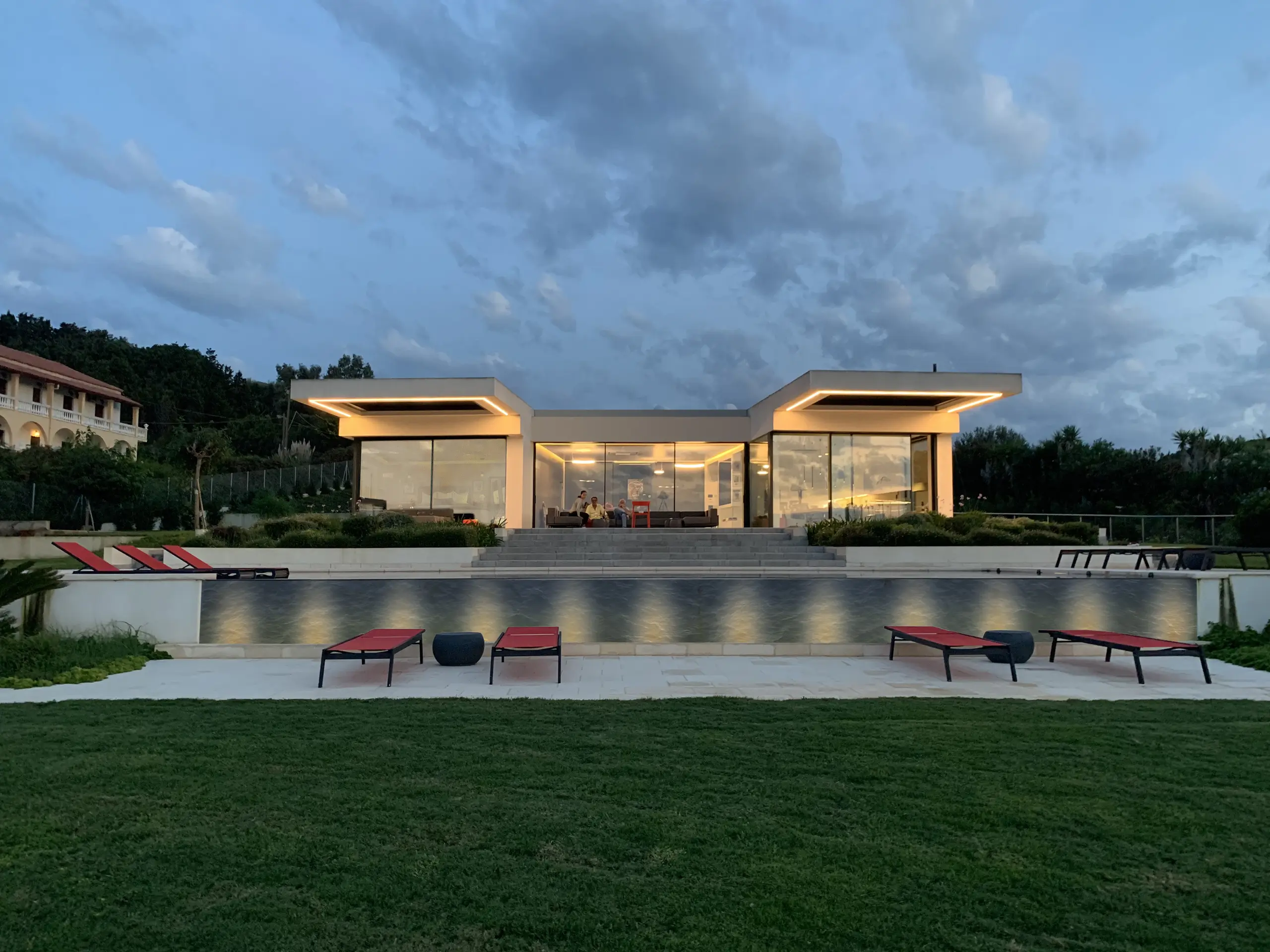Illuminated single-storey glass pavilion at dusk, fronted by a lawn and terrace lit with red and grey loungers