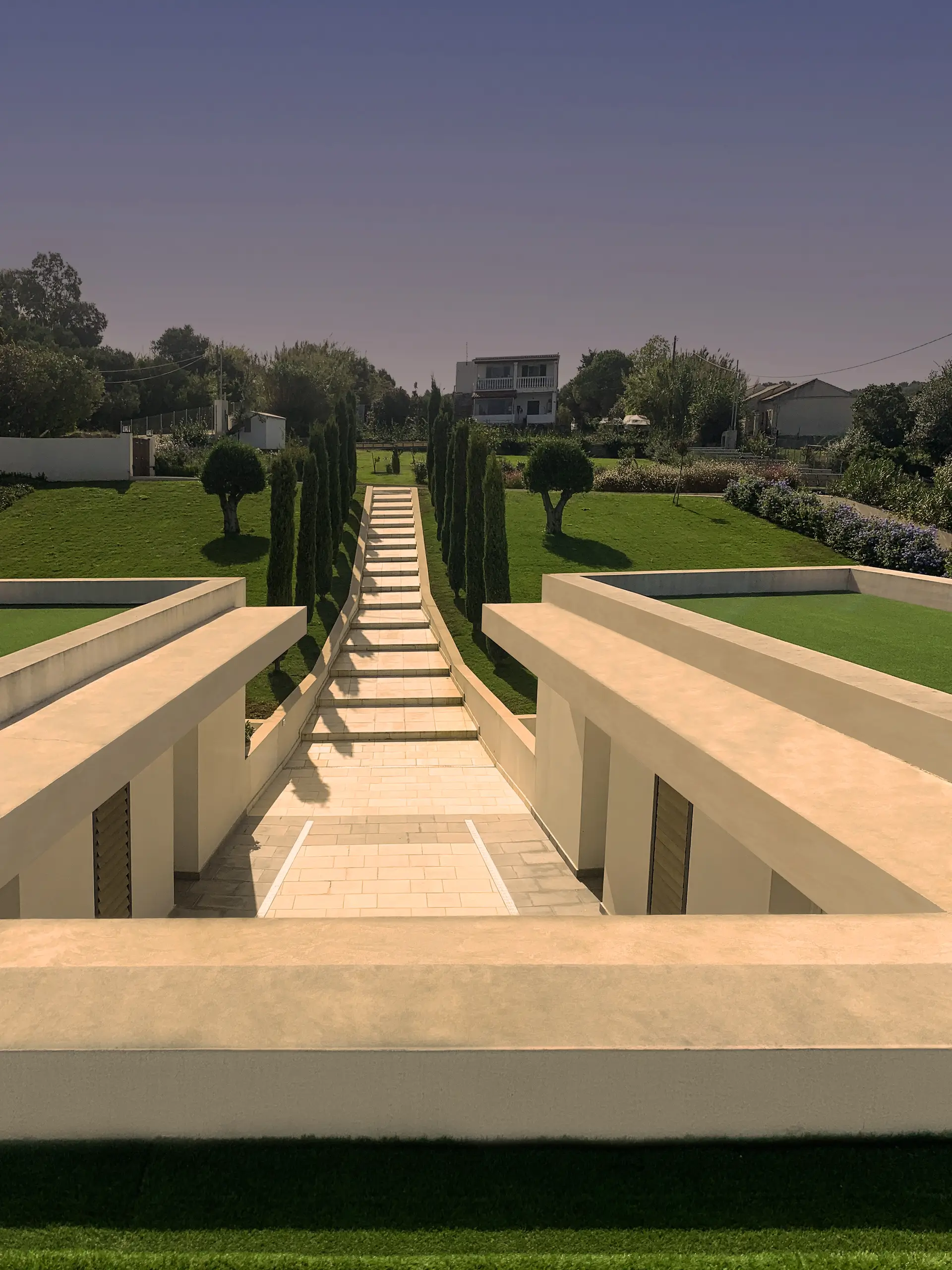 Symmetrical garden axis flanked by cypress trees, with a stepped pathway rising between low rendered walls toward the hillside