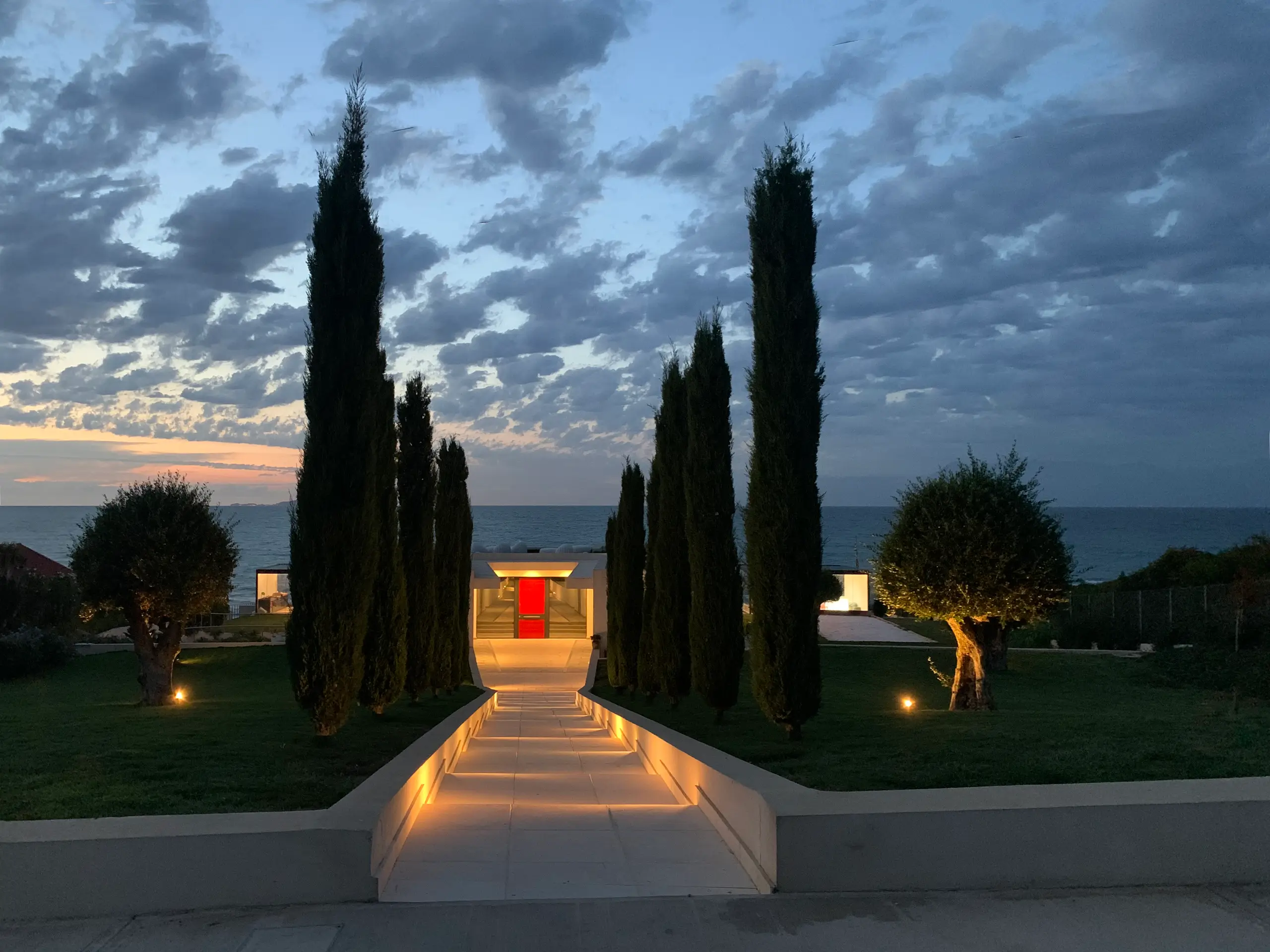 Lit walkway at dusk descending between tall cypresses toward a glowing red doorway, with the sea beyond