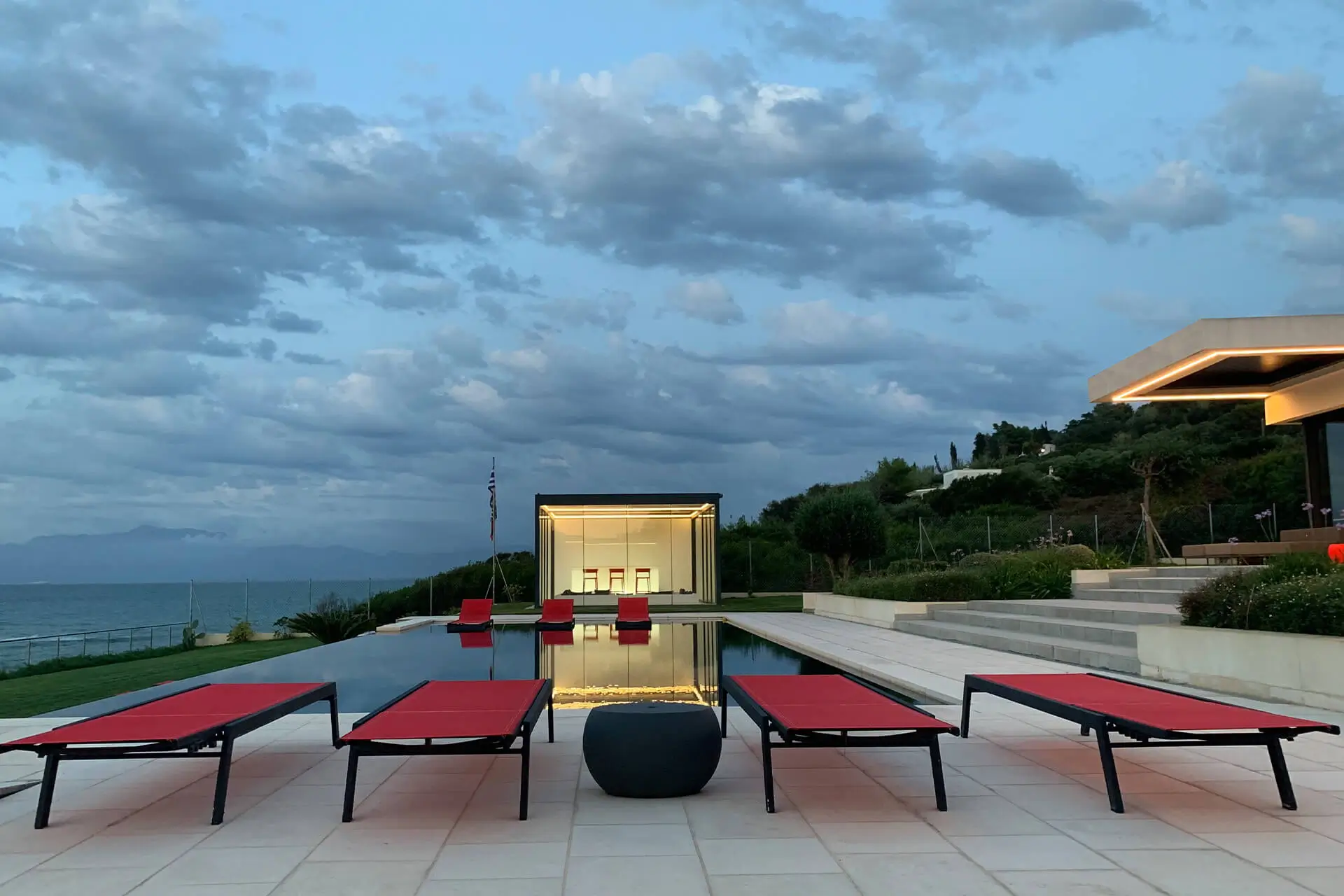 Infinity pool at dusk reflecting an illuminated glass pavilion, edged by red loungers with the Ionian Sea and distant mountains beyond